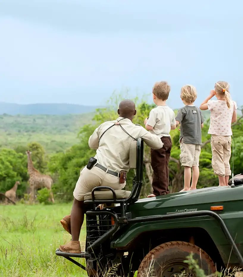 Ein Safariguide sitzt auf der Motorhaube seines Jeeps. Drei Kinder stehen neben ihm auf der Motorhaube. Sie betrachten gemeinsam Giraffen, die ein Stück entfernt vor Bäumen im hohen Gras stehen