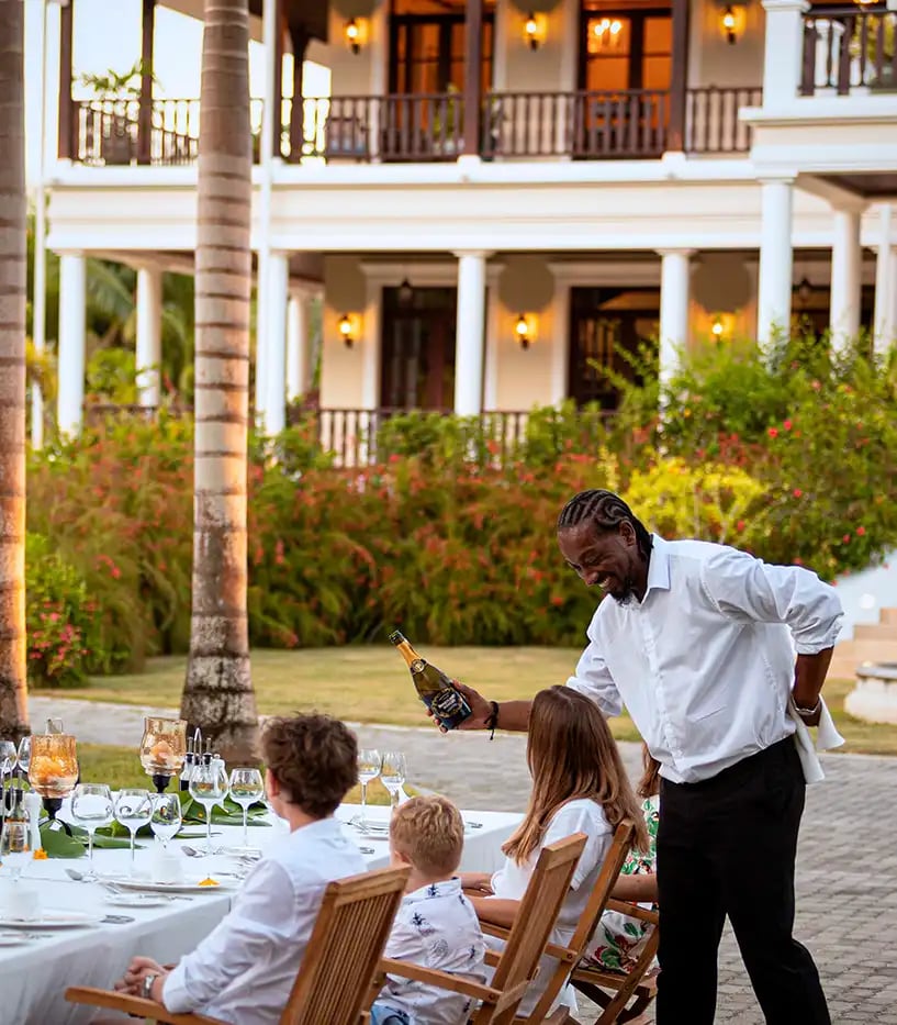 Eine Familie sitzt an einem Dinnertisch im Freien vor einer Ville. Das Servicepersonal schenkt Getränke ein.