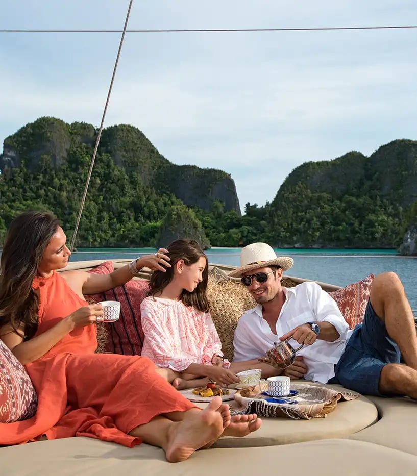 Eltern liegen mit ihrer Tochter auf einer bequemen Sitzgelegenheit auf einer Privatjacht im Meer. Im Hintergrund sind kleine Inseln zu sehen.