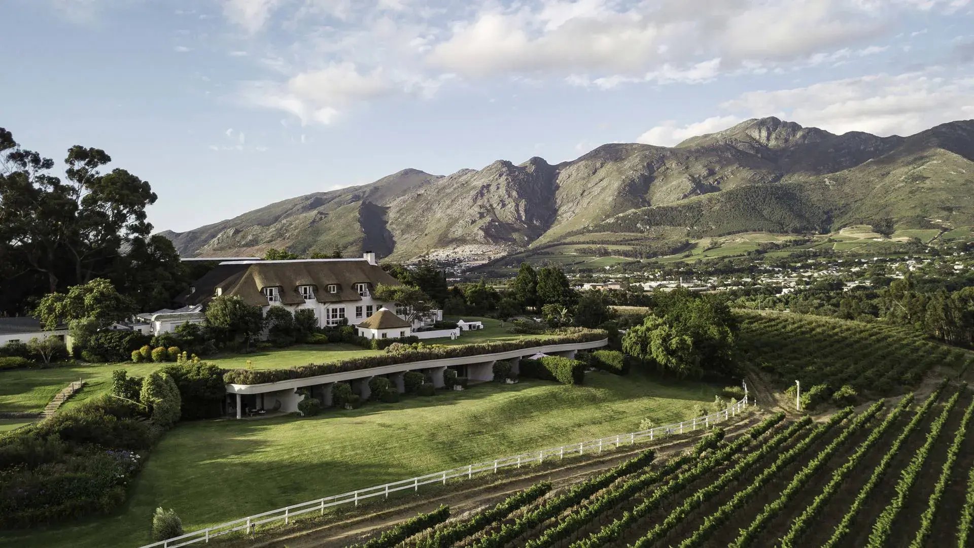 Weinberge mit Bergpanorama. Darin steht eine kleine Hotelanlage.