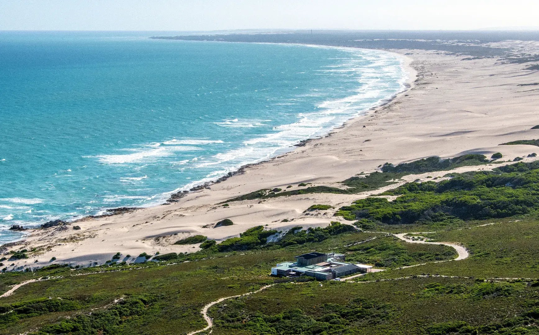 Landschaftsaufnahme einer Küste mit weiten Sandstrand, grüner Vegetation und einer Lodge, die klein zu erkennen ist.