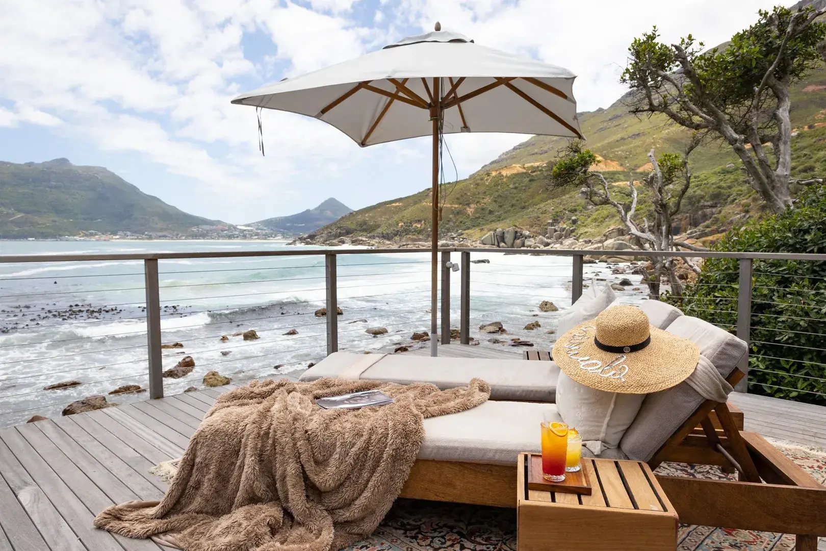 Terrasse am Strand, auf einer Liege liegt eine Decke mit Sonnenhut, ein Sonnenschirm ist aufgespannt.