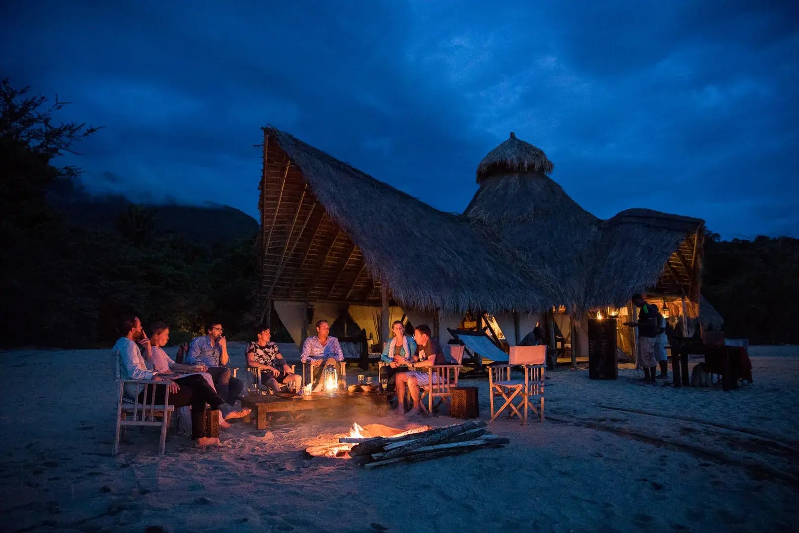 Eine Reisegruppe sitzt abends am Lagerfeuer am Strand vor einer Lodge.