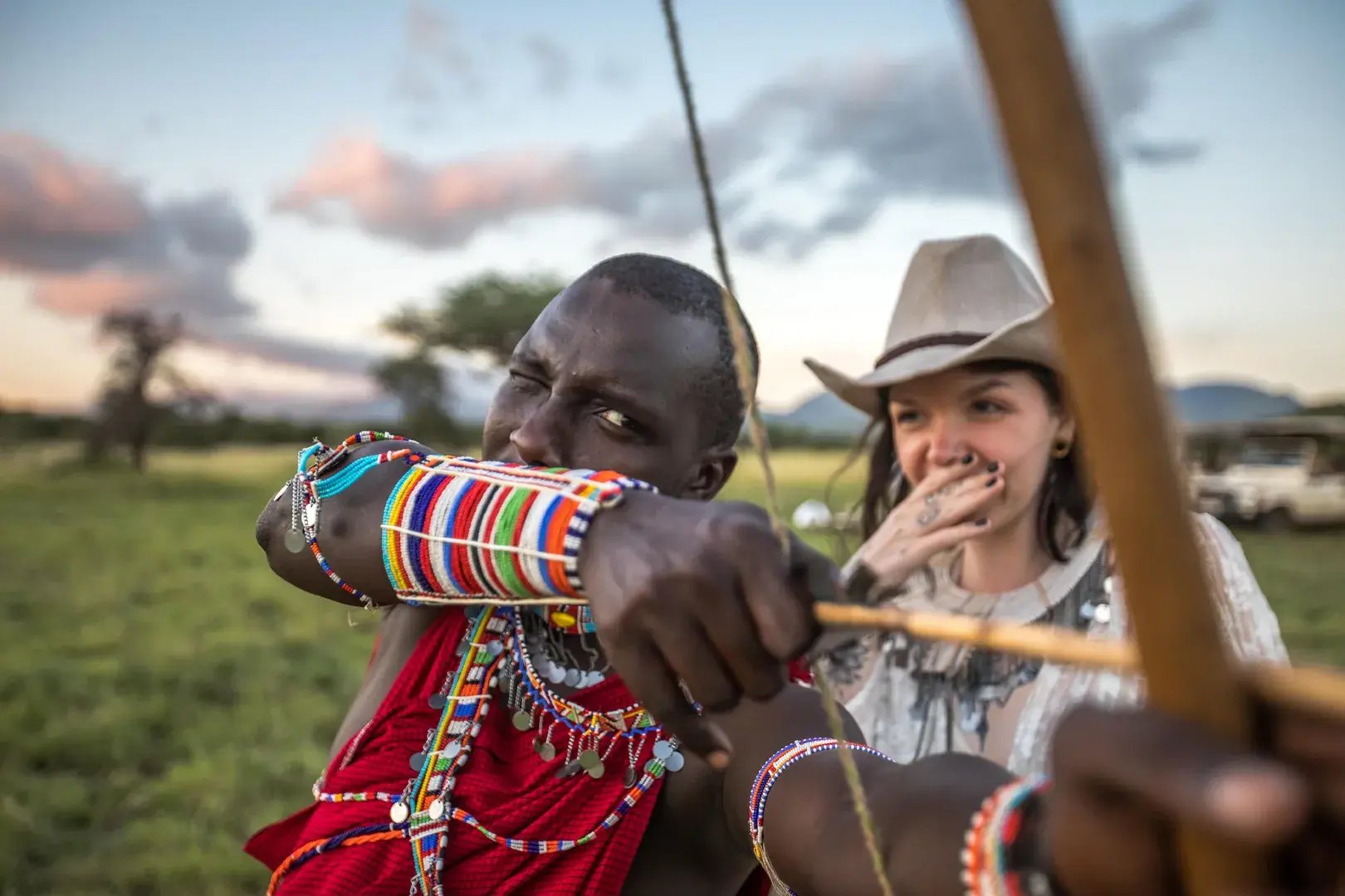 Ein Maasai-Krieger spannt den Bogen und zielt auf etwas. Hinter ihm steht eine Touristen.