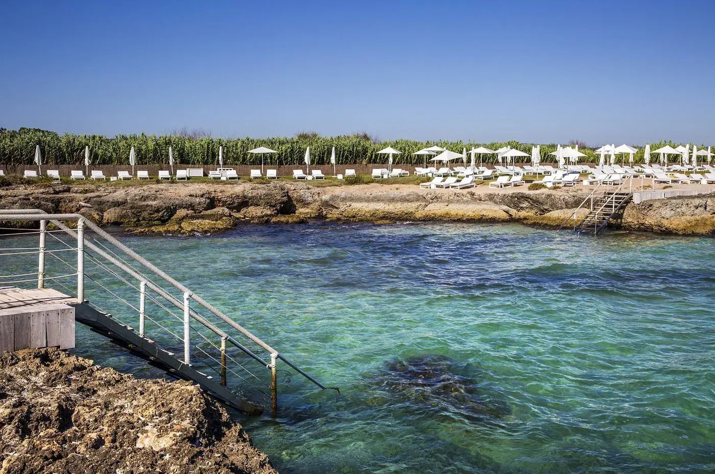 Naturpool mit Steinstrand, an dem viele Liegen und Sonnenschirme stehen.