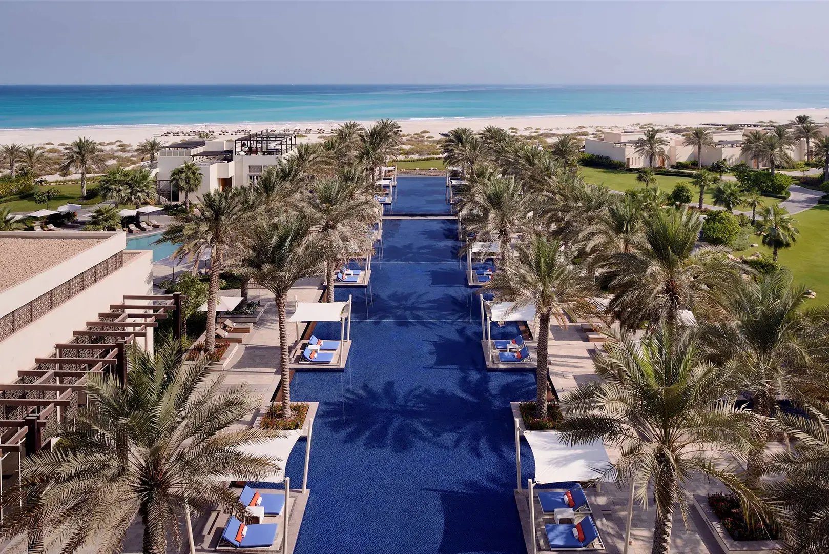 Länglicher Pool mit vielen Palmen und Liegen, im Hintergrund der Strand sowie das Meer